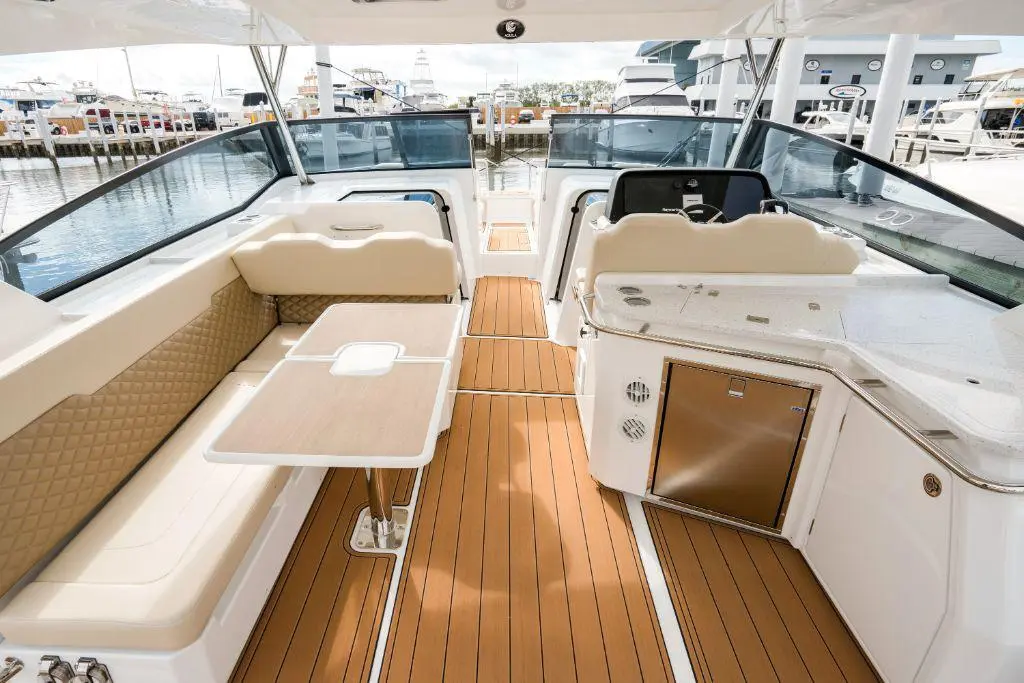A spacious yacht interior featuring tan seating, a wooden table, and modern controls, overlooking a marina with sailboats.
